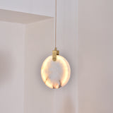 Luna LED Alabaster Disc Pendant Light 200mm in brushed brass, glowing warm white, hung by a brass hinge and cable in a light-toned room corner.