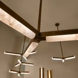 Close-up of the Elvaris LED two-tier radial chandelier, showing brushed brass copper arms with glowing alabaster bars converging at a central hub, additional fixtures visible in the background.