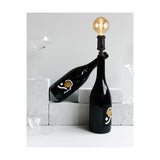 Kork E27 Table Lamp fitted to a black wine bottle neck with a globe bulb, styled beside empty glasses and pale stone blocks against a light background.