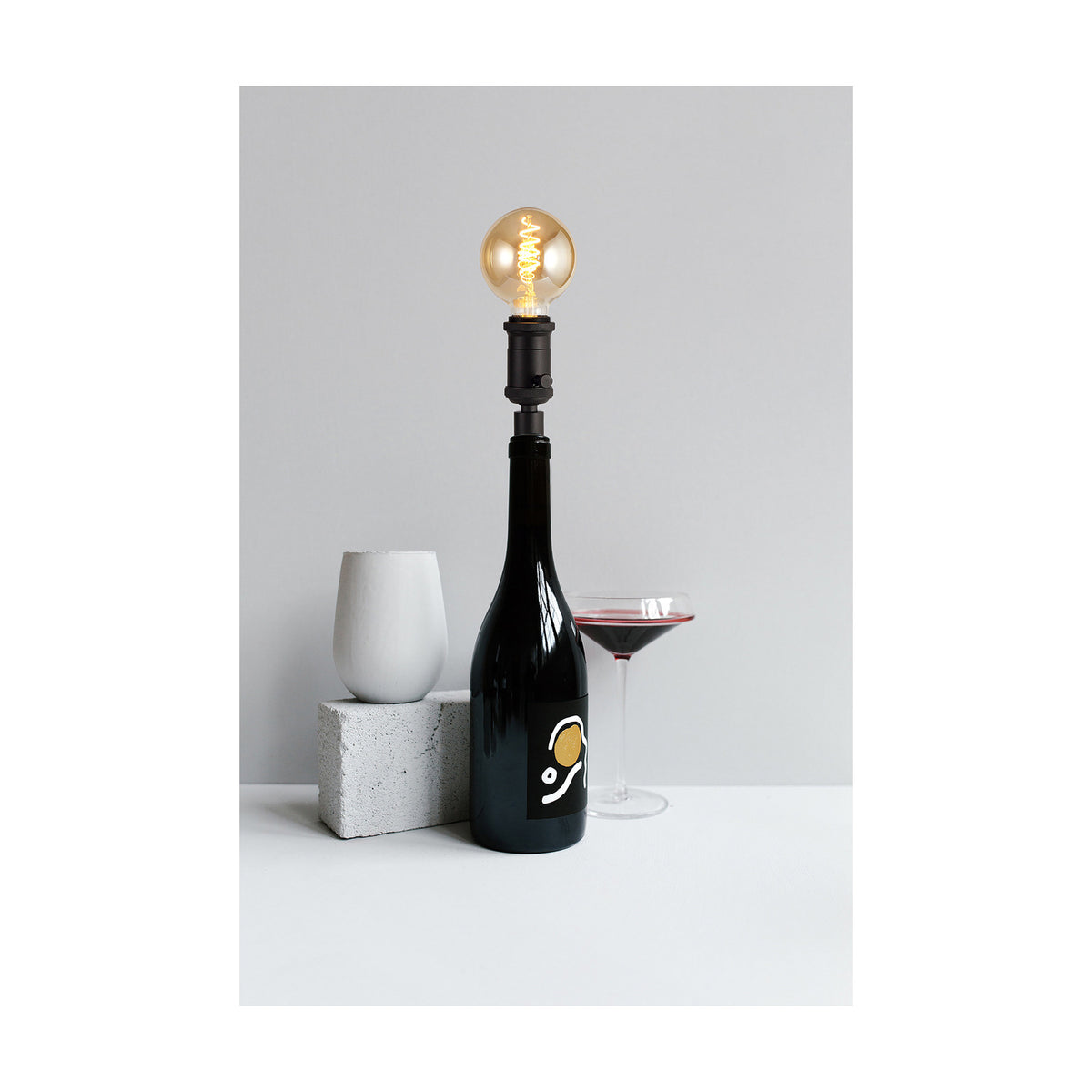 Kork E27 Table Lamp 60mm in black, iron bottle-neck fitting with rotary switch holding a globe bulb atop a dark wine bottle, styled with a wine glass and concrete accents against a grey background.