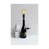 Kork E27 Table Lamp 60mm in black, iron bottle-neck fitting with rotary switch holding a globe bulb atop a dark wine bottle, styled with a wine glass and concrete accents against a grey background.