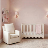 Dancer Convertible Cot Bed in cashmere finish with scalloped base, shown in a pink nursery beside a cream armchair with a folded blanket draped over the side.