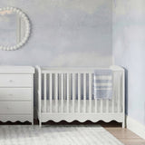 Dancer Convertible Cot Bed in white MDF with slatted sides and scalloped base, shown in a nursery beside a matching chest of drawers and round mirror.