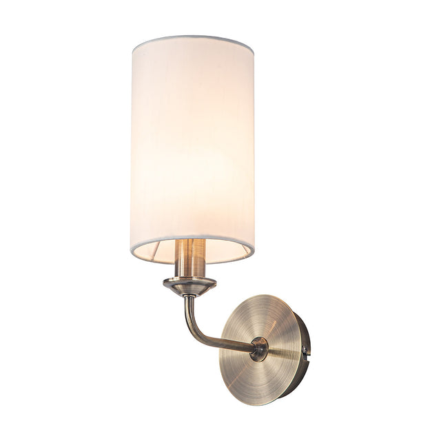 Banyan E14 wall light with antique brass curved arm and round backplate, white faux silk cylindrical shade, and rocker switch, shown on a plain white background.