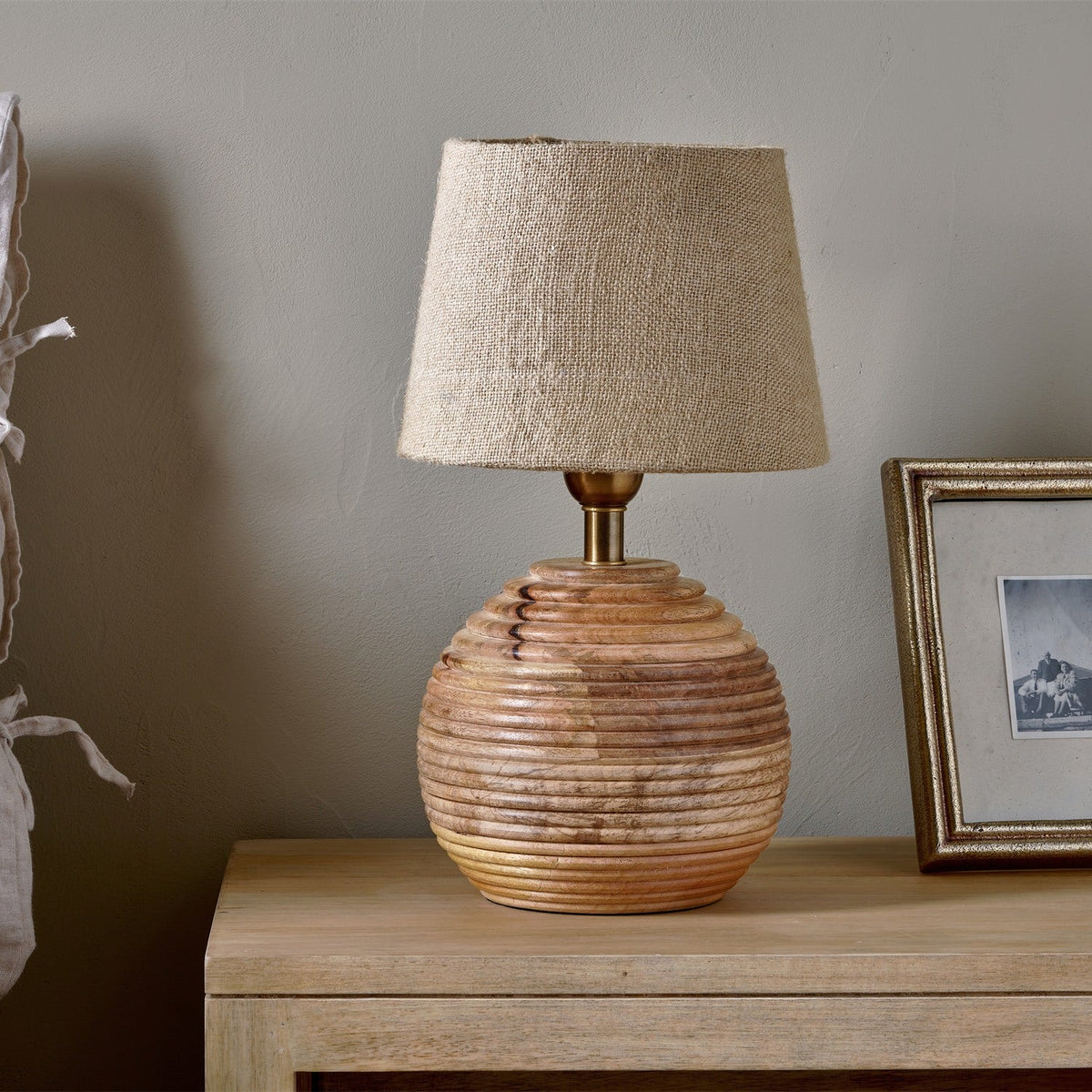 Drisana Mango Wood Bedside Table Lamp Base with ridged round base and linen shade, shown on a wooden sideboard beside a framed photo.