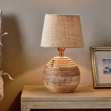Drisana Mango Wood Bedside Table Lamp Base in Natural, a rounded ribbed wood base with brass collar, shown lit with a textured shade on a wooden sideboard.