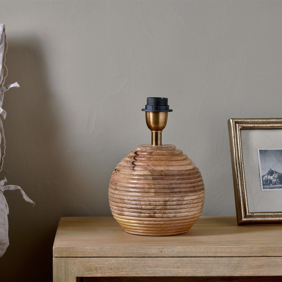 Drisana Mango Wood Bedside Table Lamp Base in Natural, a rounded ribbed wood base with brass collar and black E27 lampholder, shown on a wooden sideboard beside a framed photo.