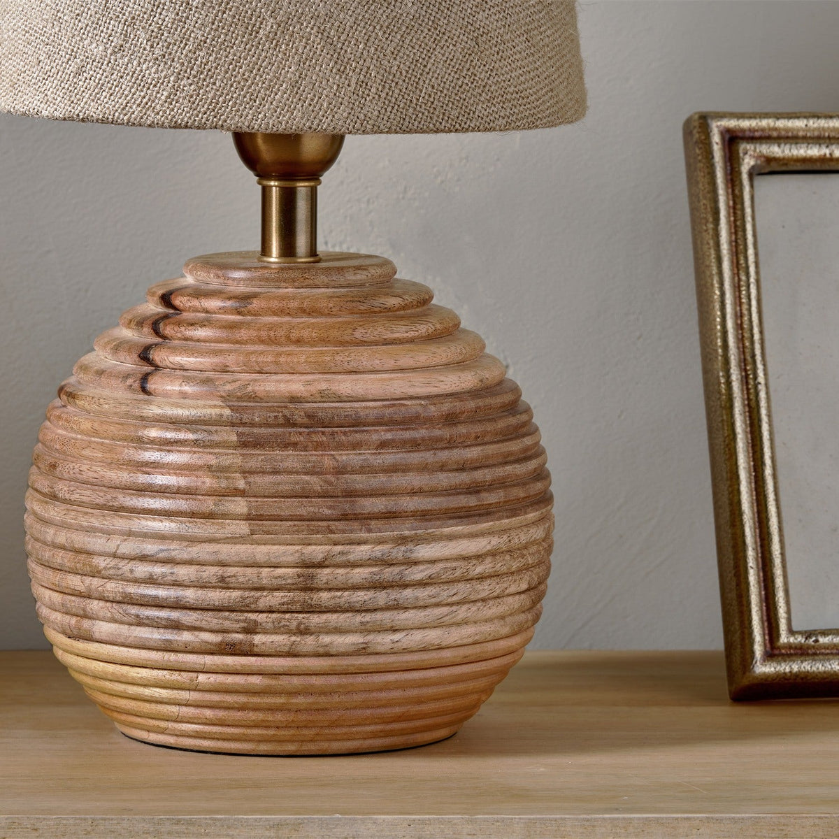 Drisana Mango Wood Bedside Table Lamp Base in Natural, a rounded ribbed wood base with grooved rings and brass collar, shown with a linen shade on a wooden surface beside a mirror frame.