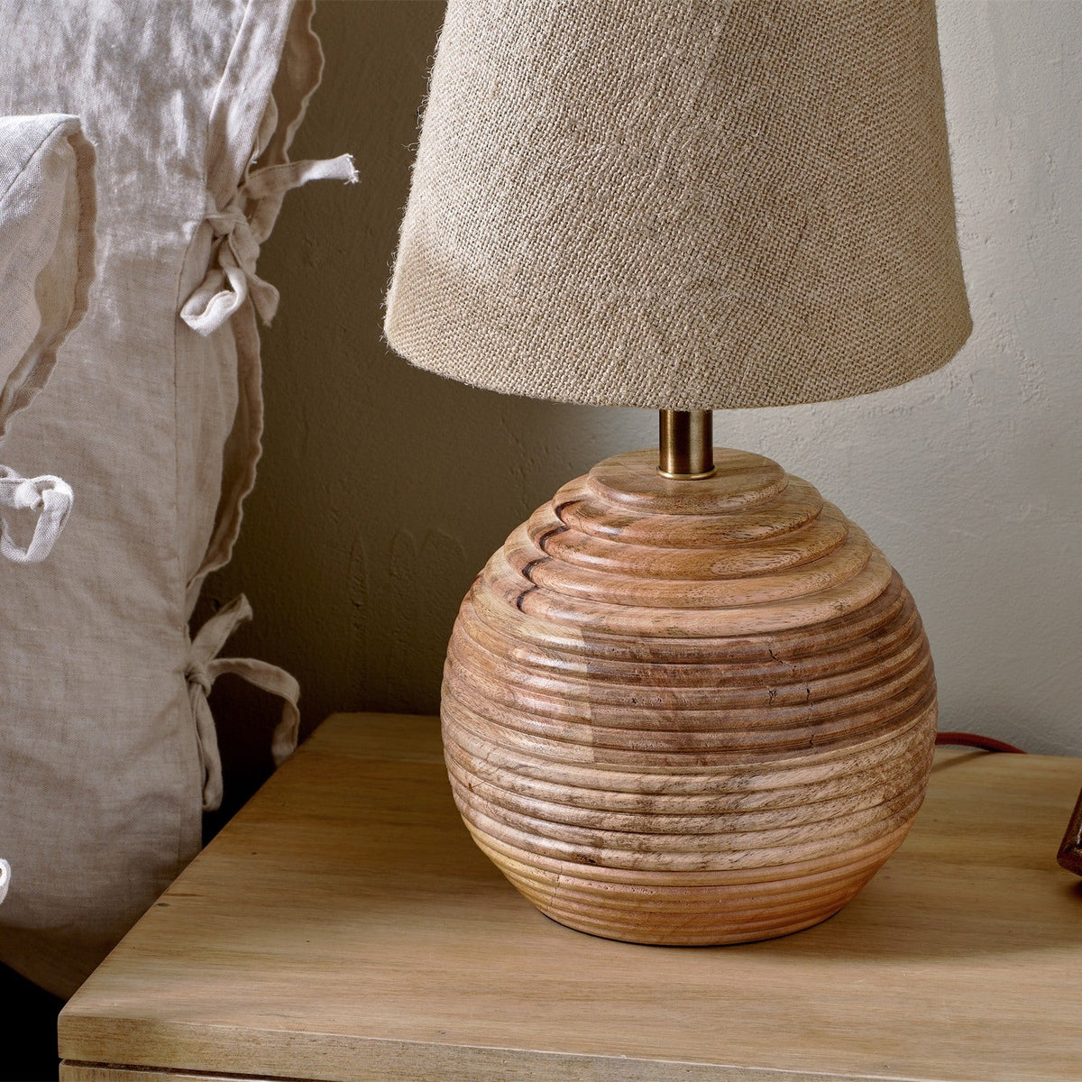 Drisana Mango Wood Bedside Table Lamp Base in natural tones, shown with a linen shade on a wooden nightstand beside layered bed linens.