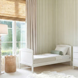 Dreamer Convertible Cot Bed in white MDF, shown as a toddler bed with panel headboard in a nursery beside striped wallpaper and a white chest of drawers.