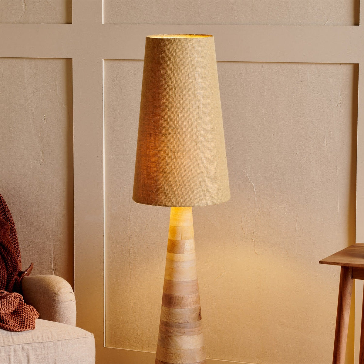 Dia Medium Jute Tall Lampshade in natural straw-coloured woven jute, tapered cone shape, shown lit atop a wooden floor lamp base in a living room setting.