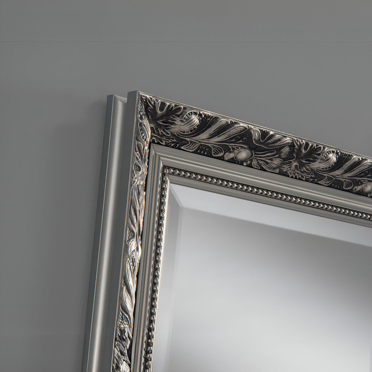 Close-up of an ornate picture frame corner featuring intricate floral and leaf patterns. The luxurious silver finish of the Dahlia Rectangle Wall Mirror beautifully contrasts against the smooth, light gray wall, adding classic elegance to the simple backdrop.