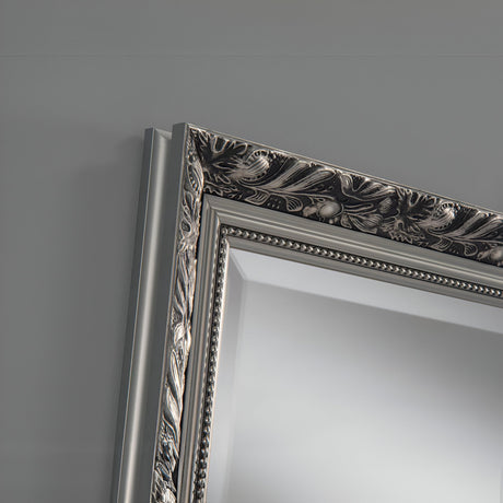 Close-up of an ornate picture frame corner featuring intricate floral and leaf patterns. The luxurious silver finish of the Dahlia Rectangle Wall Mirror beautifully contrasts against the smooth, light gray wall, adding classic elegance to the simple backdrop.