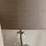 Close-up of the Embaata Small Linen Lampshade in natural, tapered drum shape with visible flax slub texture, fitted atop a clear glass lamp base with chrome gallery.