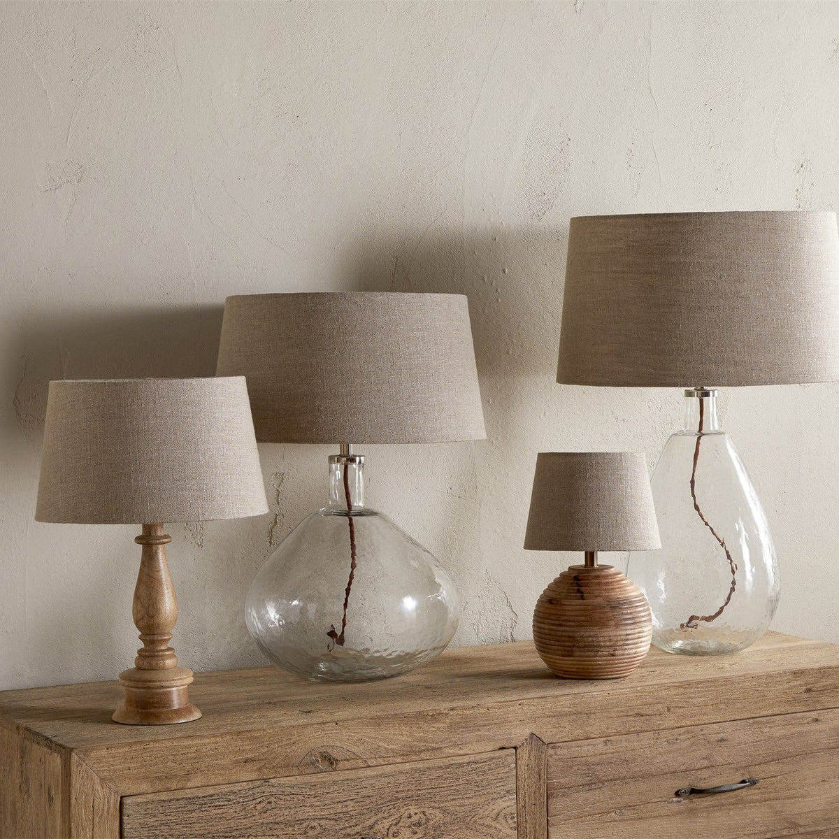 Embaata Large Linen Lampshade in natural greige shown atop four table lamps with wooden, ribbed, and clear glass bases on a wooden sideboard.