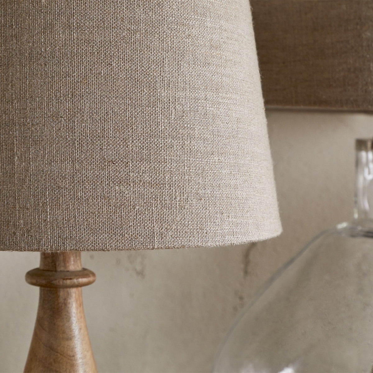 Close-up of the Embaata Large Linen Lampshade in natural greige, tapered drum shape on a turned wood lamp base, with a second shade visible behind.