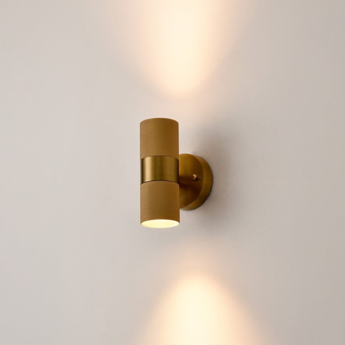 Exto Antique Brass Outdoor Wall Light with knurled cylindrical body and central band, mounted on a plain wall casting light up and down, GU10 fitting.