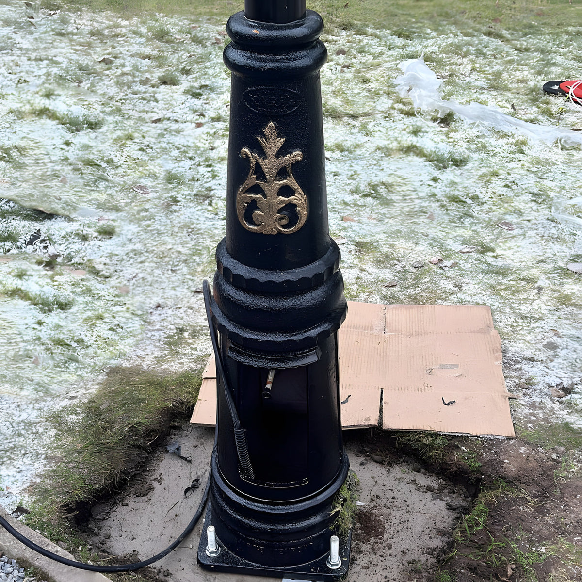 Base of a black lamp post with gold detailing mounted on a square galvanised steel foundation frame with four visible bolts, set outdoors on frosty grass.