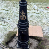 Base of a black lamp post with gold detailing mounted on a square galvanised steel foundation frame with four visible bolts, set outdoors on frosty grass.