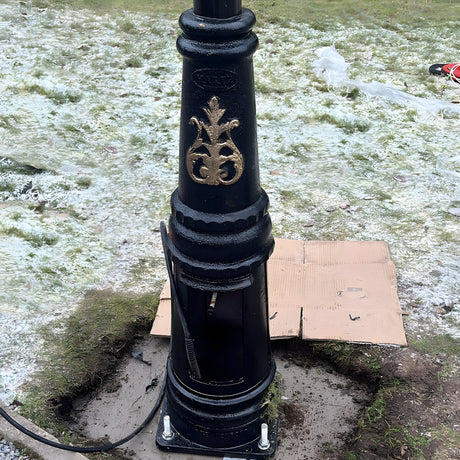 Base of a black lamp post with gold detailing mounted on a square galvanised steel foundation frame with four visible bolts, set outdoors on frosty grass.