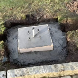 Medium Foundation Frame 150mm Square set in wet concrete outdoors, showing four galvanised steel studs and a central conduit pipe within a dug garden trench.