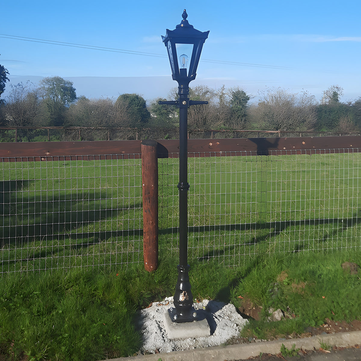 Black traditional lamp post with lantern-style top installed in a garden setting, mounted on a concrete base containing a galvanised steel 150mm square foundation frame.