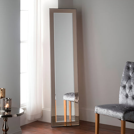 The Focus Leaner Mirror - Bronze, a sleek, rectangular full-length mirror, leans against a light gray wall. Beside it is a plush, gray tufted chair. A round side table with a lit candle is situated near a window adorned with sheer white curtains.