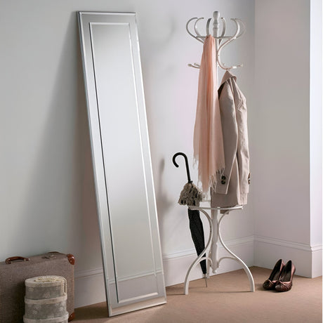 The Focus Leaner Mirror, featuring a sleek silver frame, leans against a white wall. Nearby, a white coat rack holds a beige jacket, a pink scarf, and an umbrella. A pair of brown shoes and a vintage suitcase with an additional smaller case rest on the floor.