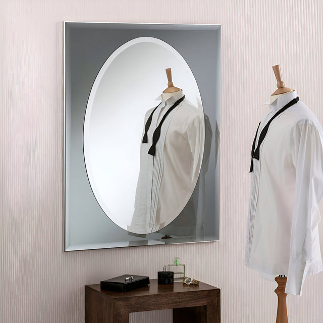 A wooden mannequin dressed in a white shirt and black tie stands in front of the Vivid Oval Wall Mirror Grey - 83x62cm. The mirror's reflection enhances the scene with a decorative touch. Beneath it, a wooden table displays a small black box and a green perfume bottle.