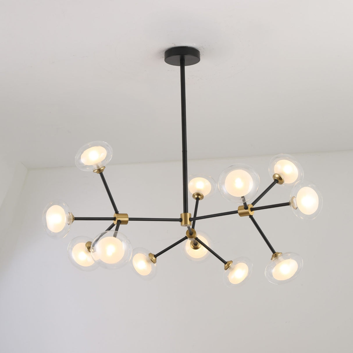 Niori Galaxy 12 Light Branching Chandelier with matt black asymmetric arms, brushed brass collars and clear glass globes, shown lit against a white ceiling.