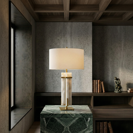 The Orinth 1 Light Framed Alabaster Column Table Lamp in Brass & Soft White with a white shade rests on a green marble pedestal in a stylish gray-walled room featuring built-in shelves, books, and a small sculpture in the background.