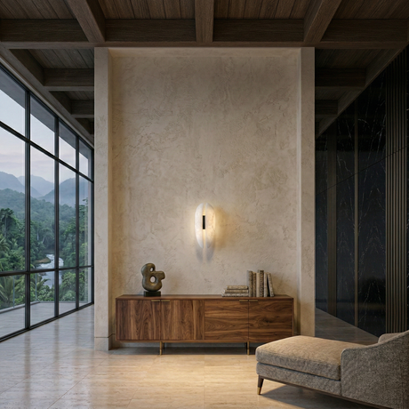 A modern interior features a large window overlooking greenery, a wooden sideboard with books and a sculpture, a textured wall with the Majorae LED Small Split Alabaster Wall Light in Soft White & Matte Black, and a gray armchair on marble flooring.