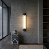 The Velisse LED Vertical Core Alabaster Wall Light in Soft White & Black casts a soft, diffused glow on a textured concrete wall beside a marble pedestal with a ceramic vase; shelves and large windows accent the minimalist space.