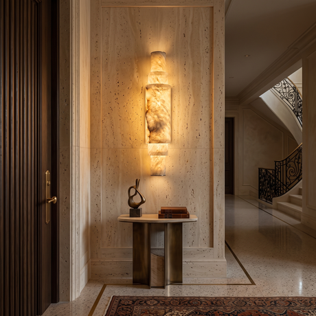 Niori Aurelia LED alabaster wall light in brushed brass, stacked cylindrical column illuminated on a travertine wall above a round bronze console table.
