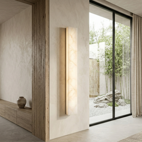A modern minimalist interior showcases the Aureline LED Extra Large Sculpted Column Alabaster Wall Light - Soft White on a textured wall, with wood accents, a ceramic vase, large glass doors, and an outdoor garden. Natural light brightens the space.