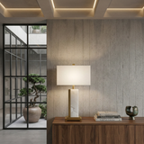 A modern interior features a wooden sideboard with the Silvarin 1 Light Sculpted Alabaster Table Lamp in Brass & Soft White, stacked books, and a sculpture. Behind, glass doors open to bonsai trees in a sunny courtyard.
