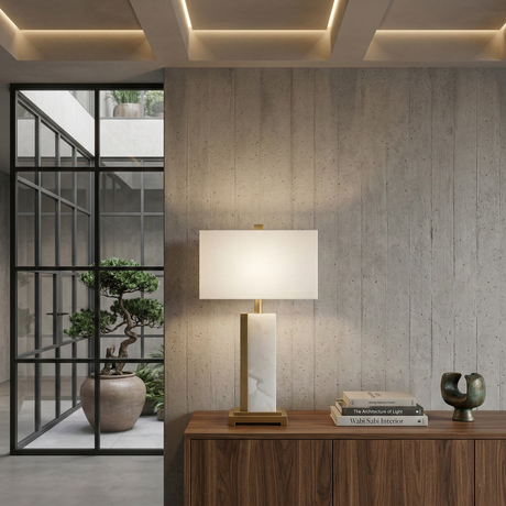 A modern interior features a wooden sideboard with the Silvarin 1 Light Sculpted Alabaster Table Lamp in Brass & Soft White, stacked books, and a sculpture. Behind, glass doors open to bonsai trees in a sunny courtyard.