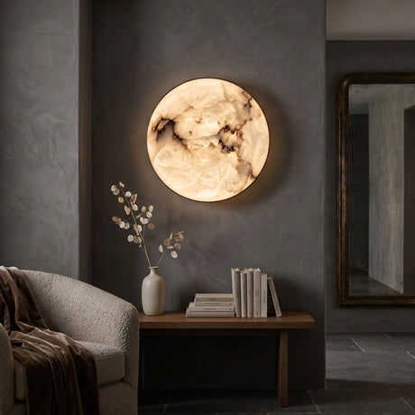 Orvani LED Medium Round Alabaster Wall Light glowing warm white on a dark wall, its natural stone diffuser lit above a wooden console in a living room setting.