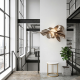 A modern loft interior with large windows, a metal wall sculpture, a Circulael 45cm Alabaster Coffee Table – Brushed Brass, a potted plant, and industrial-style railings with exposed pipes.