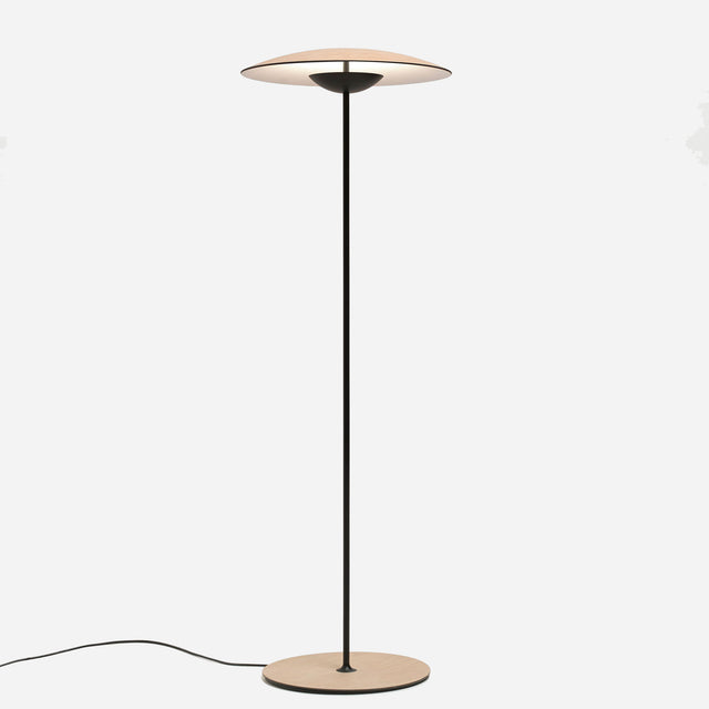 The Ginger LED Floor Lamp - Black & Oak enhances modern interiors with its sleek design, featuring a circular oak wood base and a disc-shaped beige shade. The black lamp stands elegantly against a plain white backdrop, complemented by its gracefully extending power cord from the base.