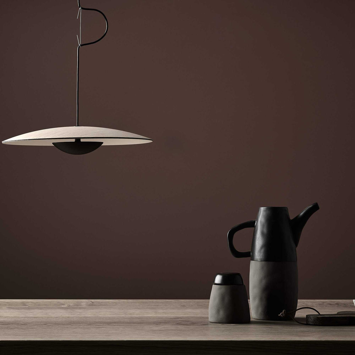The scene showcases a minimalist design with an eco-conscious touch—a sleek Ginger LED Pendant Light in Black & Oak hanging from above. Below, on a wooden surface, rests a modern black and gray teapot accompanied by a matching cup, all set against a smooth, dark brown wall.