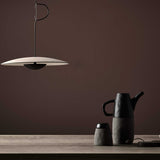 The scene showcases a minimalist design with an eco-conscious touch—a sleek Ginger LED Pendant Light in Black & Oak hanging from above. Below, on a wooden surface, rests a modern black and gray teapot accompanied by a matching cup, all set against a smooth, dark brown wall.