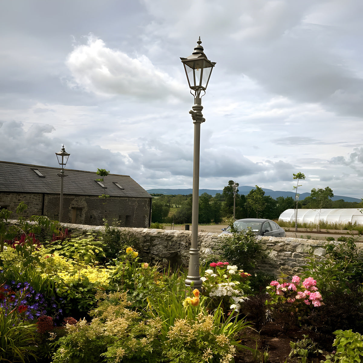 Victorian Cast Iron Lamp Post 3.5m in black, tall traditional column with tapered four-sided lantern, shown in a garden setting beside stone walls and flower beds.