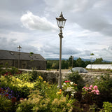 Victorian Cast Iron Lamp Post 3.5m in black, tall traditional column with tapered four-sided lantern, shown in a garden setting beside stone walls and flower beds.
