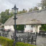 Victorian Cast Iron Lamp Post 3.5m in black with tapered four-sided lantern, shown on a stone patio beside a thatched-roof building with outdoor seating.