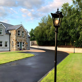 Victorian Cast Iron Lamp Post 2.6m in black with a tapered four-sided lantern head and cast finial, lit and positioned beside a driveway leading to a stone-detailed house.