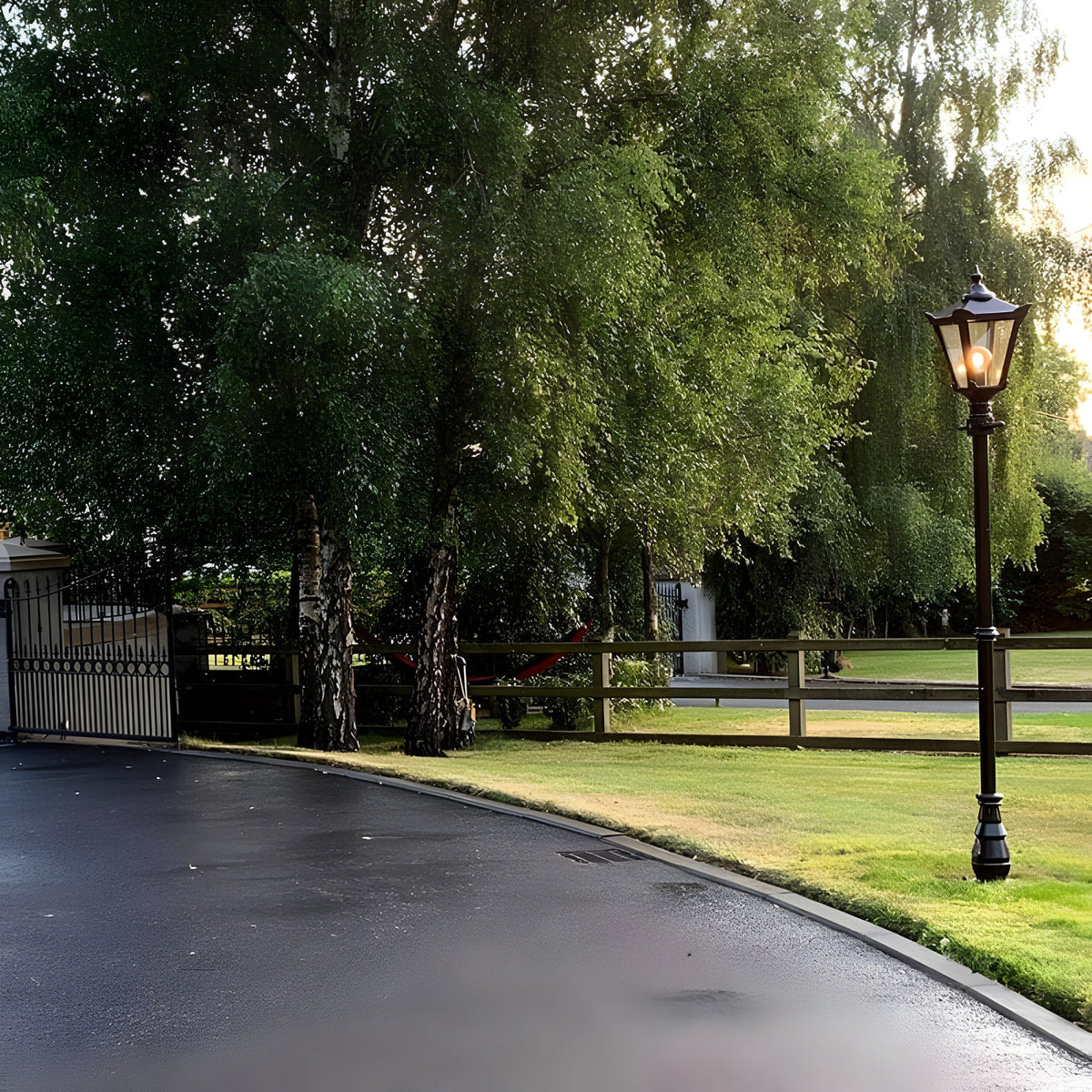 Victorian Cast Iron Lamp Post 2.6m in black, single-light lantern head with glass panels on a tapered post, shown illuminated on a lawn beside a driveway and gate.