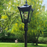 Victorian Cast Iron Lamp Post 2.3m in black with a tapered four-sided lantern head and exposed globe bulb, set on a lawn against trees with yellow blossom, a deer visible behind.