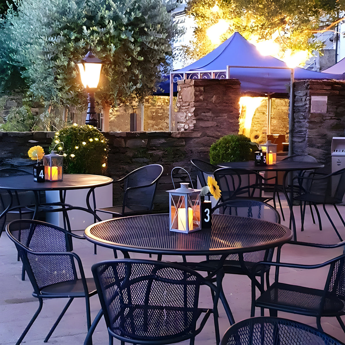Large Victorian cast iron pedestal post light in black, illuminated on a garden patio with a tapered lantern head, glowing amid outdoor dining tables at dusk.