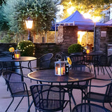 Large Victorian cast iron pedestal post light in black, illuminated on a garden patio with a tapered lantern head, glowing amid outdoor dining tables at dusk.